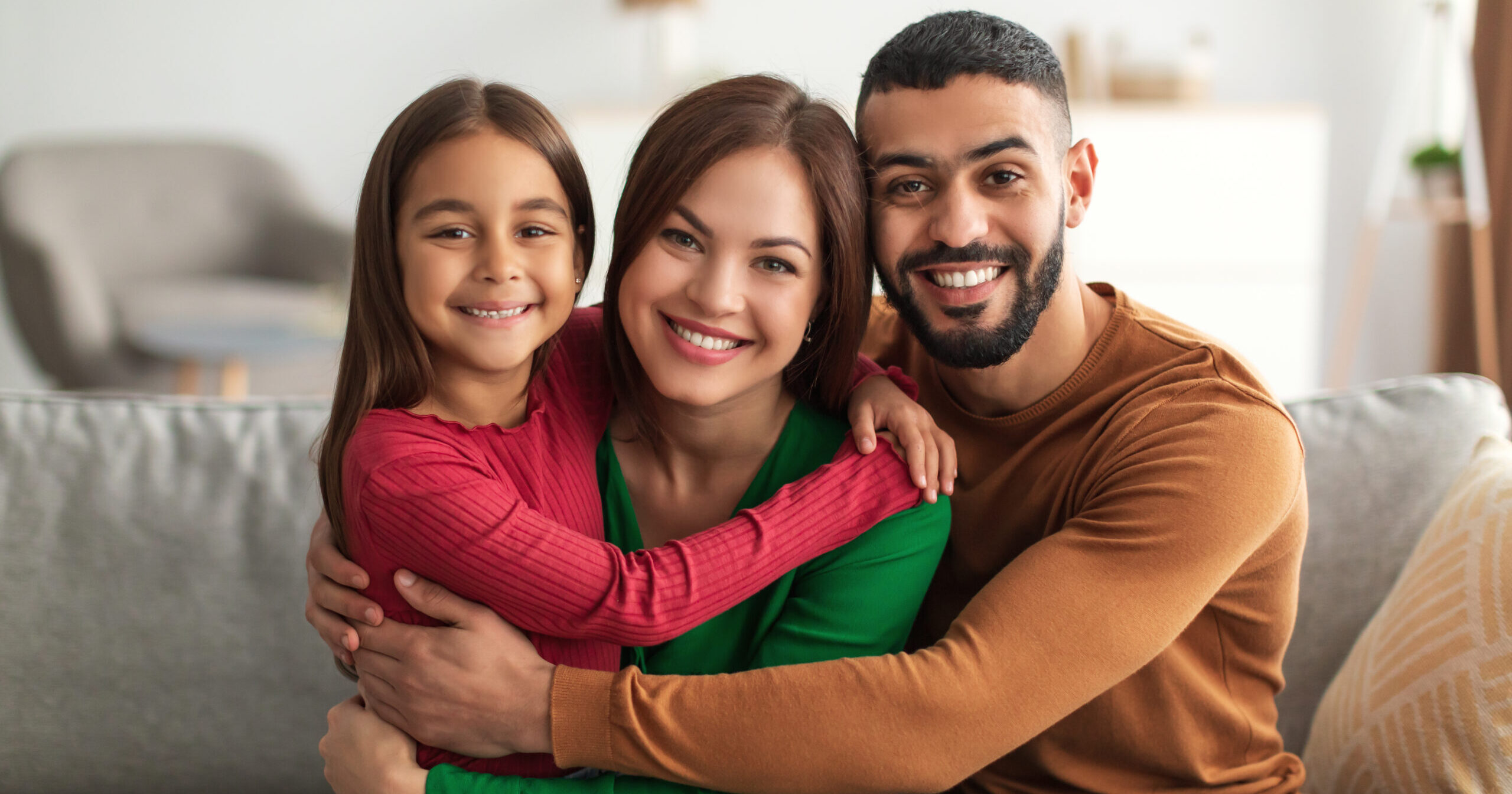 Portrait of cheerful Arabic family of three people hugging sitting on the couch at home, posing for photo and looking at camera. Smiling young bearded man embracing his loving wife and daughter